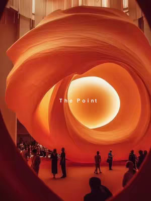 The image features a large, abstract sculpture resembling a giant orange rose or spiral rising against a backdrop of a modern interior space. In the foreground, a heart-shaped frame surrounds the main subject, framing the image. Within the sculpture's core, there is a bright, circular source of light that illuminates the interior, drawing the eye into the depths of the structure. The base of the image shows silhouettes of people in an audience, adding scale and context.