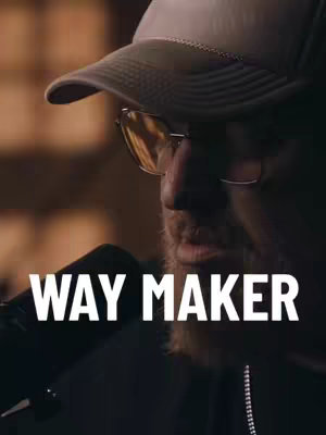 The image shows a close-up of a man wearing a baseball cap and sunglasses, holding a microphone close to his mouth. Text overlayed in white, in a bold sans-serif font, reads "WAY MAKER". The background features a blurred, warm-toned environment suggesting an indoor setting, perhaps a performance or recording space.