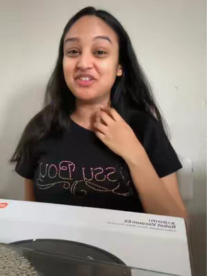 The image shows a young woman in a black t-shirt speaking to the camera. She has long, dark hair and is touching her neck with her hand. In front of her is a box with text on it, and a glimpse of a robot vacuum cleaner is visible.