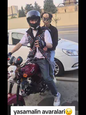 Two young men are on a motorcycle. The man in the front, who appears to be riding the bike, is wearing a black helmet, a vest, and blue jeans. His face is partially obscured. The other man has a Momo-like mask. The scene is set against a blurred background with a white car, buildings, and trees.