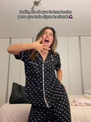 A woman in polka-dot pajamas is captured in a full-body shot against a bedroom backdrop. She is mid-action, with her mouth wide open, and one hand raised towards her face, suggesting an emphatic expression. The background includes a bed with a pink sheet and a wardrobe.
