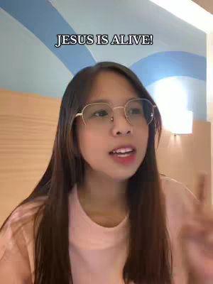 A young woman with long dark hair and glasses faces the camera and speaks. Behind her is a light wall. The overlay text at the top of the image says 'JESUS IS ALIVE!'