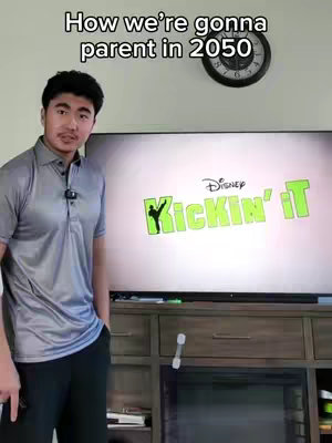 A young man stands in front of a large TV screen displaying the Disney show "Kickin' It." He's wearing a gray polo shirt and black pants. Above the TV and behind the man, a clock hangs on the wall.