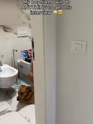 A person is sitting on a toilet with a laptop. A dog is curled up on the floor looking towards the camera. The image is shot from a doorway.