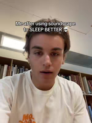 A young man with brown hair and a white t-shirt is looking at the camera with a relaxed expression. Behind him is a bookshelf filled with books.