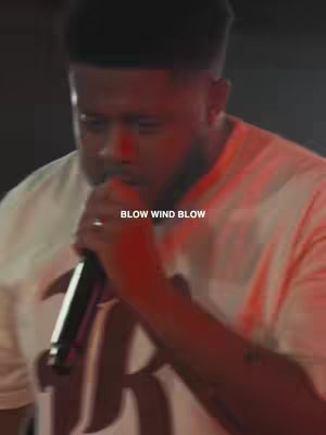 The cover image shows a man with short, dark hair singing into a microphone. The lighting is dim, with red hues, and the text overlay "BLOW WIND BLOW" is displayed in white, centered at the bottom.