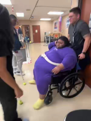A woman in a purple outfit and wheelchair is being pushed by a man. There are other people in the background and the setting is an indoor area, likely a rehabilitation center with tennis balls placed at the feet of the woman and a person using a walker.