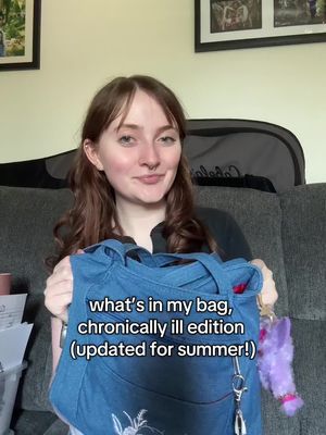 A woman with light skin and reddish-brown wavy hair is smiling and holding a blue tote bag. The bag has a white floral design. The text overlay says 'what's in my bag, chronically ill edition (updated for summer!)'. The background has a grey sofa, a guitar case, and framed pictures.