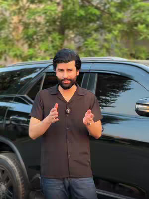 A man in a black shirt stands in front of a black car, gesturing with both hands in a forward motion.