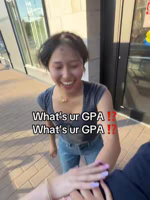 The image shows a young woman outdoors with a surprised look. She is wearing a dark blue t-shirt and jeans with a gold necklace. Her hands are up and out, and another person's hand is visible on her right. The background is a slightly blurred view of a building.