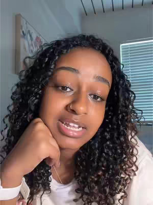 A close-up shot of a young Black woman with curly hair, looking directly at the camera with a concerned expression. She rests her chin on her left hand, and her eyes appear to be watery. The background is a blurred interior of a brightly lit room.