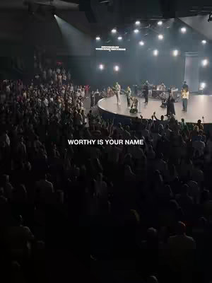 A wide shot of a large venue, likely a church or auditorium, filled with a crowd. A raised platform at the center stage hosts a band, illuminated by spotlights, creating a visual focus. The audience is seen from a slightly elevated perspective.