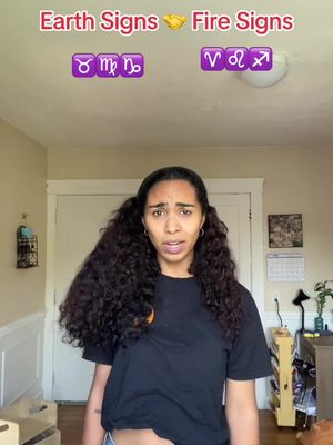 A woman with dark curly hair is the main focus, framed from the chest up. She is in a room with plain walls and a closed door. The words "Earth Signs" and "Fire Signs" are written above, along with corresponding zodiac symbols.