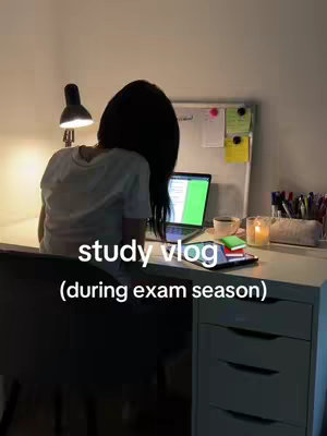 A person with long hair sits at a desk, seen from behind. They are working on a laptop that has a green screen. The desk is cluttered with study materials including books, a cup, and a candle. The scene is illuminated by a desk lamp and soft ambient light.