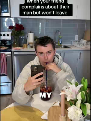 A young man sits at a table in what appears to be a kitchen, holding a smartphone and looking upwards. The background shows a kitchen scene with red roses.
