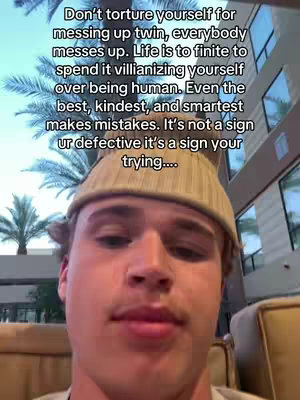 A close-up shot of a young man with fair skin and short, light brown hair, wearing a light brown beanie. He's looking directly at the camera with a neutral expression. The background shows hints of a building interior, with a blurred view of windows and architectural details. The overall composition is well-lit, with a focus on the man's face and upper body.