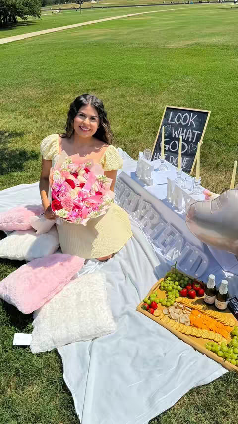 A woman sits on a white blanket in a grassy area, holding a bouquet of flowers. In front, a picnic table with food and decorations is set up. A sign that says, "Look What God Has Done" is on the table.