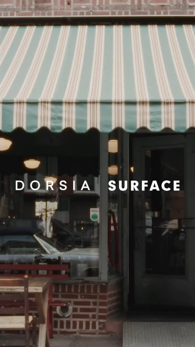 Image of the exterior of a restaurant. A green and white striped awning shades the entrance. The storefront has windows and a dark door. Text overlayed on the image reads: "DORSIA SURFACE". Partially visible through the windows is a reflection of the interior of a car, and tables are set up in front of the restaurant.