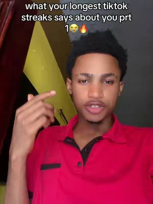 A young man with short, dark hair and medium skin tone faces the camera. He wears a red polo shirt and a black undershirt. The background is a mix of yellow and dark tones. The man is speaking and gesturing with his right hand, which is pointing upwards. Text overlays at the top, in white, includes the words "what your longest tiktok streaks says about you prt", along with a number, and two emojis.