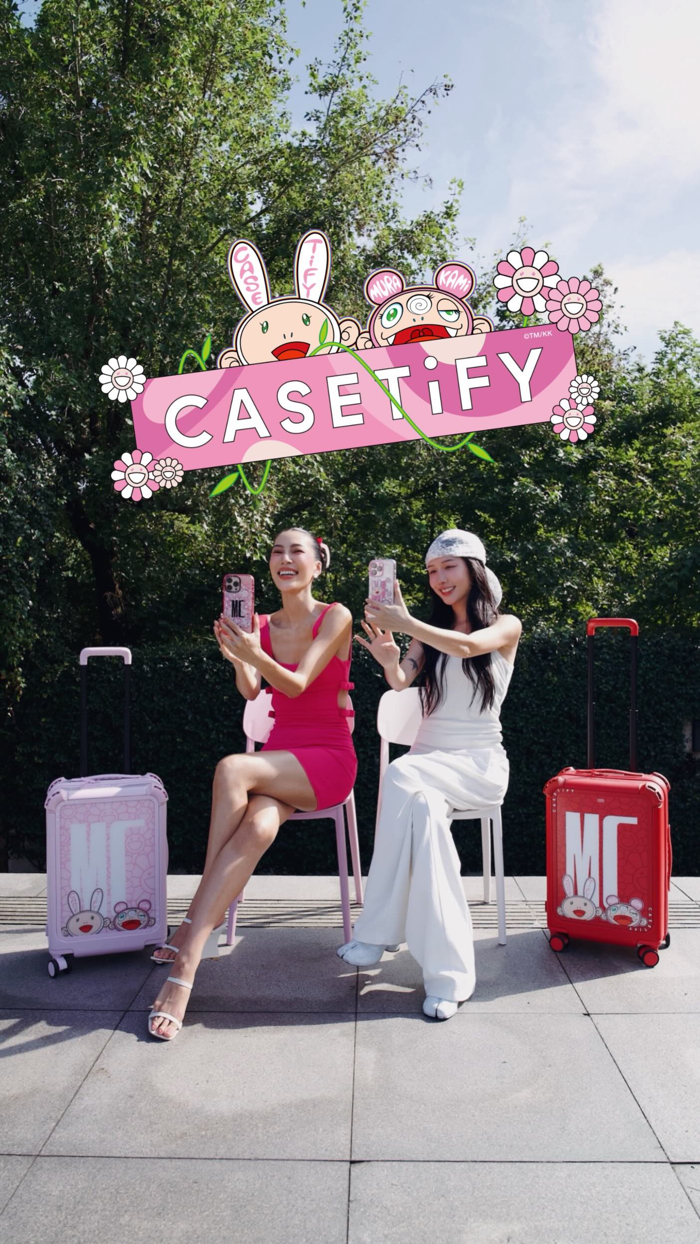 The cover image features two women, one in a bright pink dress and the other in a white outfit, each holding a smartphone and smiling, posing next to matching travel suitcases with cartoon characters. The background includes green foliage under a blue sky.
