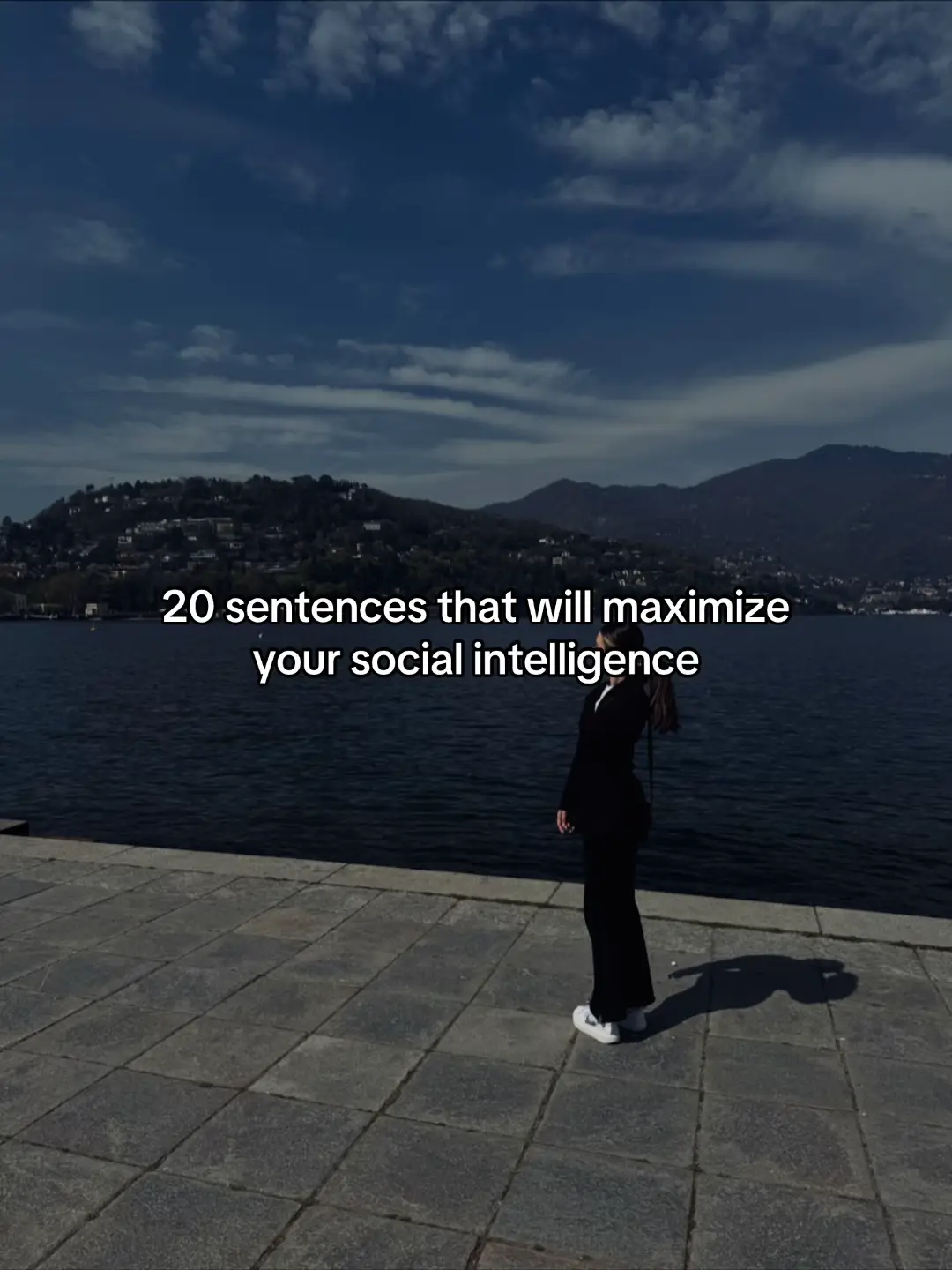 A woman in a black suit stands on a stone walkway next to a body of water. The background features a blue sky with puffy clouds and mountains in the distance. The text overlay reads: '20 sentences that will maximize your social intelligence'.