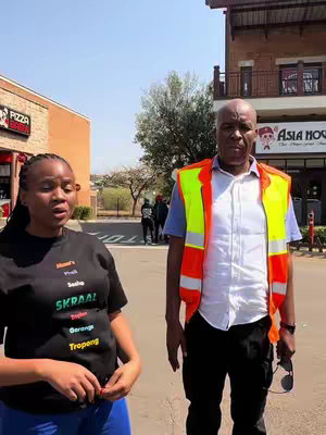 The cover image shows two people, a woman on the left and a man on the right, standing on a street. The woman is wearing a black t-shirt with text and is looking towards the camera. The man is wearing a uniform. In the background, there are buildings and other people. The scene seems to be lit by sunlight, suggesting it is daytime.