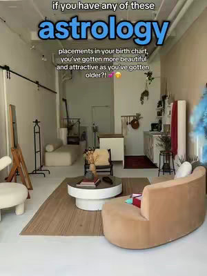 An interior shot of a modern living space with white walls, a mix of furniture, and a hardwood floor with a rug. In the foreground, there's a curved beige sofa and a round coffee table. The setting suggests it's a studio or a well-decorated apartment. The image has a clean, minimalist aesthetic.