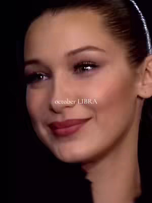 A close-up shot of a woman with light skin and dark hair. The woman is Bella Hadid, with her face taking up most of the frame. There is a text overlay with the words "october LIBRA" in a simple font. The image has a dark background with neutral skin tones and dark eye makeup. Her lips are a deep red color, and she is slightly smiling.