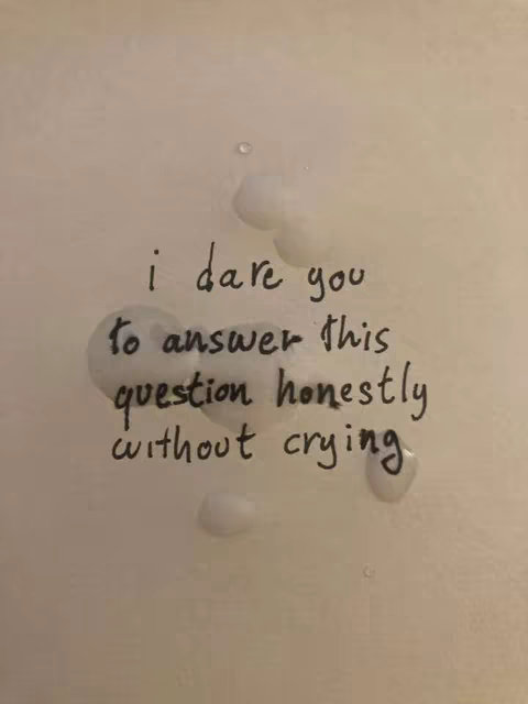 A close-up image shows a white surface, likely a wall or paper, upon which is written a question in black ink. The question, 'i dare you to answer this question honestly without crying' is surrounded by several large, irregular shapes of water, creating a visual effect suggesting tears or moisture. The text is handwritten, slightly uneven, which gives it a personal and emotional tone.