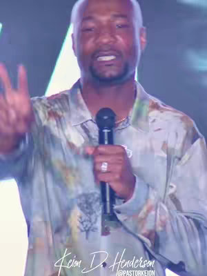 A mid-shot shows a Black man with a bald head holding a black microphone. He's wearing a light-colored, patterned shirt and a silver ring. The background is blurred, with hints of a stage setting, including lighting. The man is speaking, with his hand raised, gesturing at the audience. Text at the bottom states his name and social media handle.