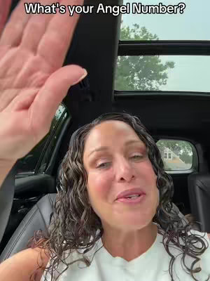 A woman with long curly hair is sitting in a car, looking at the camera with a hand raised. The background includes the car's interior and a view outside the sunroof.