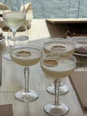 A still life shot featuring four coupe glasses filled with a pale yellow cocktail, each garnished with a dried citrus wheel. The glasses are arranged on a light-colored surface. One champagne flute and a standard drinking glass filled with water and a napkin are in the background, soft focus, along with a view of the horizon through a window. The overall composition suggests a cocktail setting.
