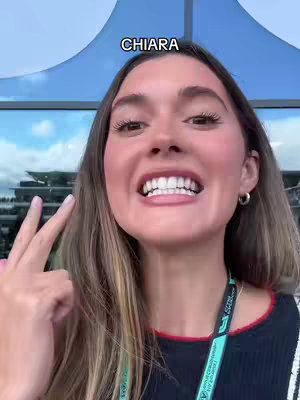 A selfie-style shot of a young woman smiling and gesturing with her fingers. The background is a blurred outdoor scene with a blue awning. The woman has long brown hair, is wearing a dark top, and has a Qatar Airways lanyard around her neck. The text overlay reads 'CHIARA'.
