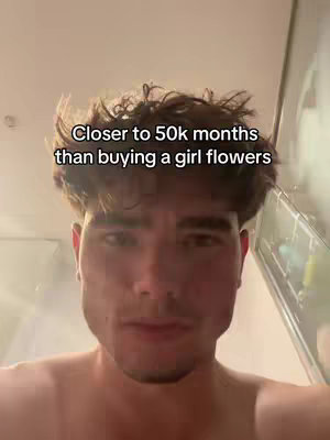 A selfie-style shot of a young man with dark hair, facing the camera. The background is a blurred setting, likely indoors. He has a neutral expression. Text overlayed on the image reads, 'Closer to 50k months than buying a girl flowers'.