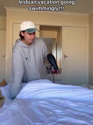 A person wearing a baseball cap and hoodie stands in a bedroom and appears to be blow-drying a bed sheet with a hair dryer attached to an electrical outlet with a red cord.