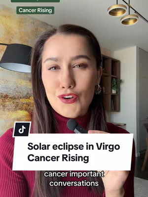 A woman with dark hair and red lipstick is the focus, and a microphone is held close to her mouth. Overlaid text on the image announces the topic of the video, a 'Solar eclipse in Virgo Cancer Rising'. The woman wears a dark red turtleneck, and neutral lighting provides a sense of professionalism. The background consists of decorative items that add context to the content as well as text related the video content.