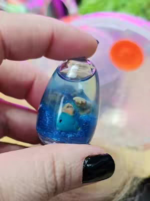 A close-up shot of a hand holding a small, clear, teardrop-shaped container. Inside, a blue liquid is filled with small, blue glitter, and a small blue dinosaur figurine. The hand has long nails painted black. The background is blurred with out-of-focus objects.