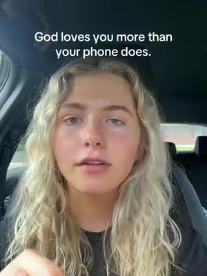 A woman with long, wavy blonde hair is centered in the frame, looking towards the camera with a neutral expression. The background suggests the interior of a car, with a partially visible side window to the left. Above the woman's head, the text overlay reads: God loves you more than your phone does.