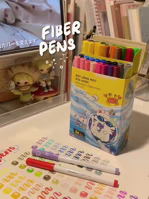 The image showcases a collection of colorful fiber pens neatly arranged in an open box. The pens are neatly grouped and presented, alongside a red and a purple pen, a sheet of paper filled with strokes from different pens colors. A monitor with a picture is visible in the background, behind the pen box. A bright, well-lit scene, suggesting creativity.