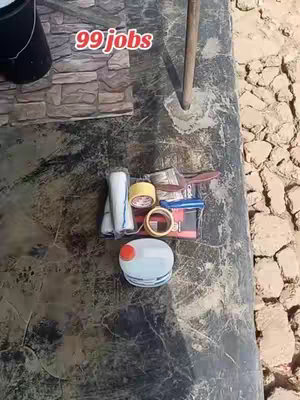 An overhead shot features a black, textured surface filled with various hardware supplies and a white container with a red top. To the left, there is a dark bucket partially visible, and to the right, a rocky background. The text '99 jobs' is placed above the objects.