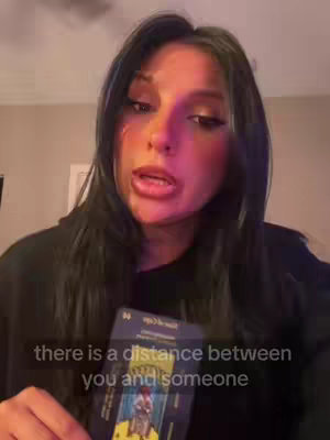 A woman with long black hair and dark makeup looks at the camera with a neutral expression, lips slightly parted. She holds a tarot card in front of her, with overlaid text at the bottom.