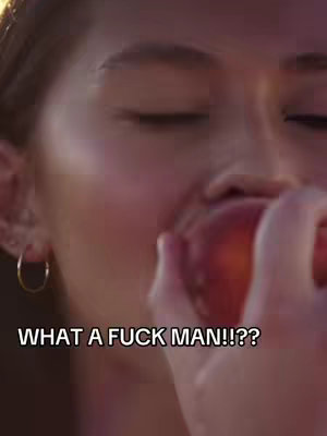 A close-up shot of a person's face, with the mouth open as if biting into a peach. Their eyes are closed, and a hand holds the peach close to their mouth. The background is softly lit.
