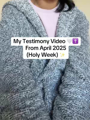 A person is seen from the chest up, wrapped in a large, gray, fluffy textured blanket or sweater with a hood up. The person is wearing a light pink shirt underneath. In the center, overlaid on top of the image, is a white rounded rectangle with black text. The text reads 'My Testimony Video', followed by a heart icon, a cross icon, 'From April 2025', and then '(Holy Week)'. The background appears to be plain.