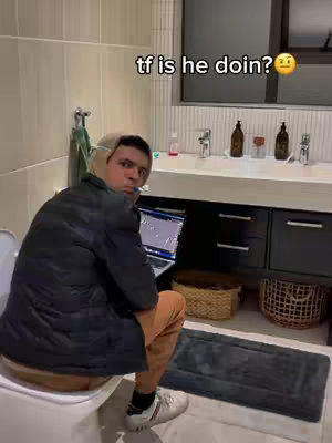 A man wearing a jacket, beanie, and sneakers, sits on a toilet, with a laptop on his lap, in a bathroom setting.
