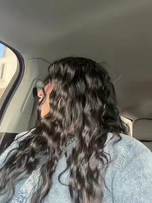 A person is filmed from a slightly angled perspective inside a car. The focus is on their curly black hair and the back of their denim jacket. They are looking out the car window.