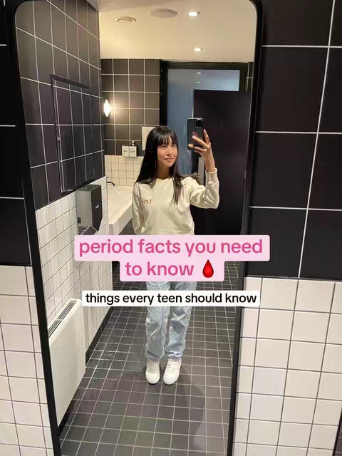 A young woman stands in a tiled bathroom, holding a phone up to take a selfie in front of a mirror. Text overlays appear on the image.
