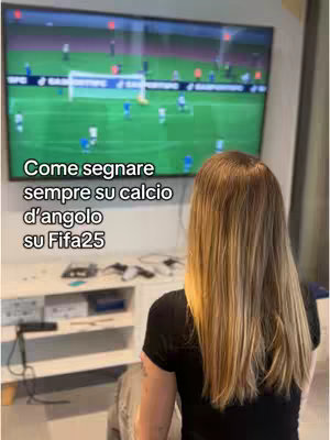 A person with long blonde hair is seen from the back, sitting and looking at a television screen. The screen displays a gameplay of a soccer match, likely from a video game, with players and the field visible.