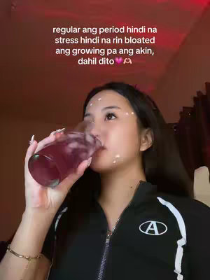 A young woman is shown from the chest up, slightly off-center, drinking from a glass indoors. The background is a blurred dark red or maroon color, suggesting a dimly lit setting. She is wearing a black zip-up jacket with a white trim around the top and the letter "A" in a white oval on the chest. The woman has long dark hair and some white dots on her face. In her hands, she is holding a glass filled with a dark pink liquid and ice cubes, tilting it towards her mouth and drinking. Her facial expression is neutral. Text is overlaid on the image in white.