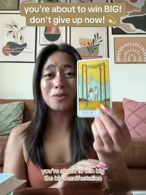 A woman with long dark hair sits in front of a backdrop of artwork and a brown couch, holding a tarot card. The cover has text overlayed, with more text at the bottom.