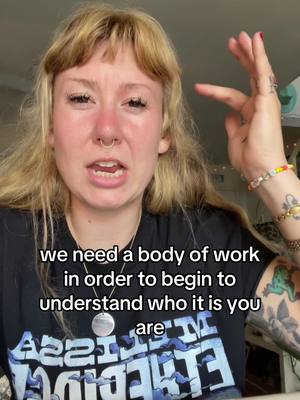 A medium shot shows a woman with long blonde hair, bangs, a nose ring, and several tattoos on her arm. She is speaking directly to the camera in a serious manner, with text overlaid across the lower portion of the image. The text is in white font against a blurred background. In the background a portion of a room is visible with the corner of a window visible and some objects on a shelf.